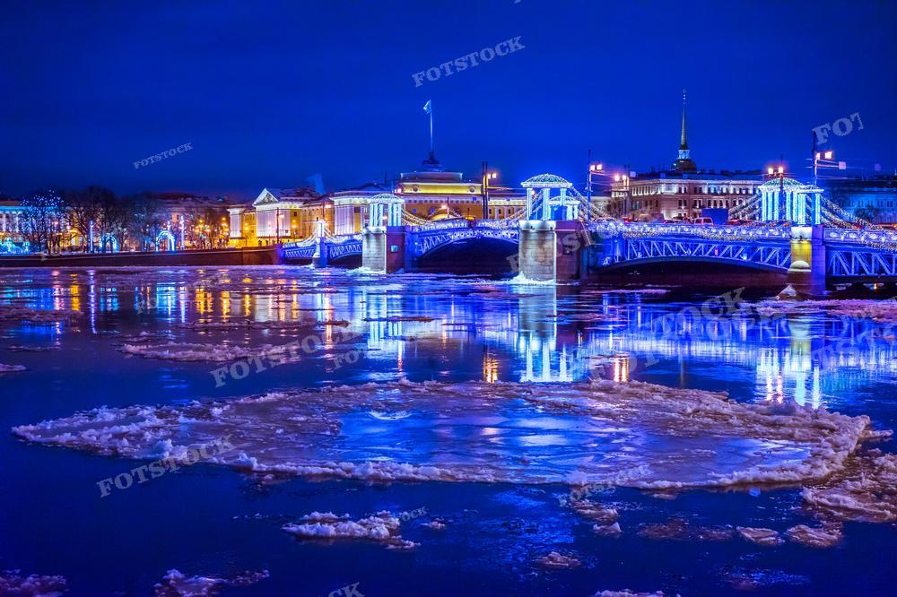 На фотографии изображён зимний вечерний вид Санкт-Петербурга. Мы видим заснеженную реку Неву и освещённый Дворцовый мост, украшенный праздничной иллюминацией. На переднем плане в реке видны льдины, что указывает на холодное время года. Фотография может использоваться в туристических буклетах, для иллюстрации статей о Петербурге или как фон для зимних праздников. Это изображение может быть полезно для дизайнеров, туристических агентств, историков и любителей городских пейзажей.